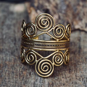 May include: A gold-toned cuff bracelet featuring a detailed pattern of swirling spirals and decorative lines. The bracelet has a crown-like top and is set against a rustic wooden surface. The jewellery is a statement piece.