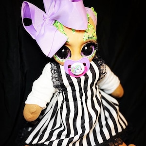 May include: A handmade doll with green and black painted details, wearing a black and white striped dress with a purple bow and a pacifier.