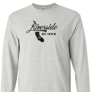 May include: A light gray long-sleeve shirt with the word "Riverside" in a cursive font. Below the word is a black silhouette of California with "CA" inside, and the text "EST. 1870" with a star.