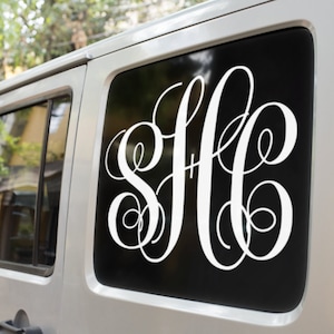 May include: White monogram decal on a black background, affixed to a vehicle window. The decal features three stylized, intertwined letters. The vehicle is silver, and the background includes trees and foliage.