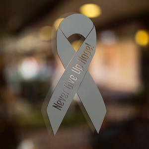 Peut inclure: Ruban gris clair avec l'inscription "Never Give Up Hope!" en police serif. Le ruban est un symbole de soutien et de sensibilisation, souvent utilisé pour représenter diverses causes. L'arrière-plan est flou, avec des tons chauds.