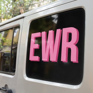 Può includere: Un veicolo argentato con un finestrino nero che mostra le lettere rosa "EWR". Le lettere hanno un effetto 3D e il veicolo è parcheggiato all'aperto con alberi sullo sfondo.