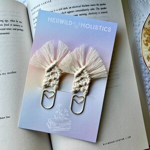 May include: Two white macrame bookmark tassels with gold heart-shaped clips. The bookmarks are on a card with a gradient background and the text "HERWILD HOLISTICS".