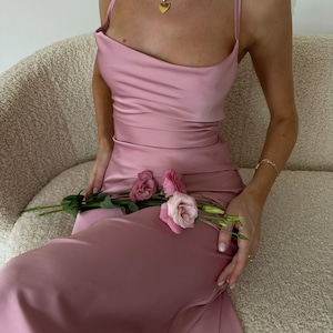 May include: A woman wearing a pink satin dress with a cowl neck and thin straps. She is sitting on a white fluffy couch and holding a bouquet of pink flowers.