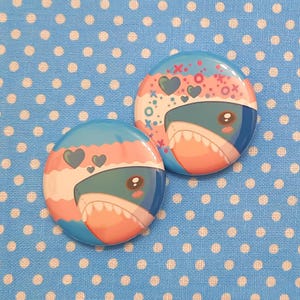 May include: Two round button badges featuring a cartoon shark design. The shark is blue, green, and pink with a white mouth and teeth. The background of the badges is blue and pink with white hearts and x's.