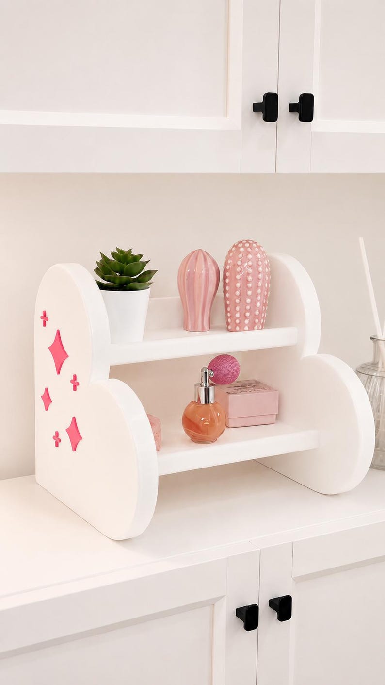 Pode incluir: Uma prateleira de madeira branca de dois n&iacute;veis com laterais arredondadas, apresentando recortes em forma de diamante e estrela cor-de-rosa. A prateleira superior cont&eacute;m uma pequena suculenta verde e dois objetos decorativos.
