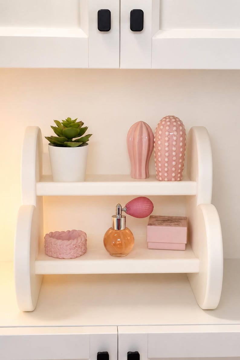 Pode incluir: Uma prateleira de madeira branca de dois n&iacute;veis com uma suculenta verde num vaso branco, um cacto de cer&acirc;mica rosa canelado, um cacto de cer&acirc;mica rosa com pontos, um anel tran&ccedil;ado rosa, um frasco de perfume de vidro e uma caixa rosa.