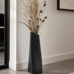Könnte beinhalten: Eine hohe, schwarze, moderne Vase mit geometrischem Design, die getrocknete Ziergräser und Samenstände enthält. Die Vase steht auf einem neutralfarbenen Boden und schafft eine minimalistische Ästhetik. Die Vase ist etwa 61 cm hoch.