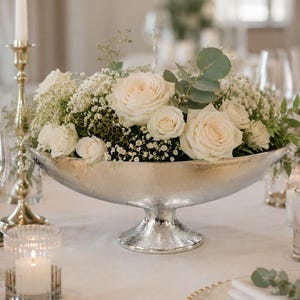 Puede incluir: Un cuenco de pedestal de metal plateado lleno de un arreglo floral de rosas color crema, paniculata blanca y hojas de eucalipto verdes. El cuenco presenta un acabado martillado texturizado y se asienta sobre una base circular.