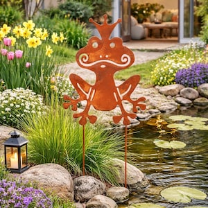 May include: A rusty metal frog garden ornament with a crown, positioned by a pond. The frog features white detailing and is set amongst flowers, rocks, and foliage. A lit lantern is also visible.
