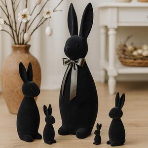 Può includere: Una collezione di statuette di conigli neri in velluto di varie dimensioni, disposte su un pavimento in legno. Il coniglio più grande ha un nastro grigio. Decorazioni pasquali con vaso, rami e uova appese.