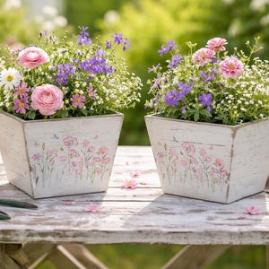 Könnte beinhalten: Zwei weiße, quadratische Holzpflanzgefäße gefüllt mit bunten Blumen, darunter rosa Rosen und lila Blüten. Die Pflanzgefäße haben eine Used-Optik und florale Designs. Die Szene spielt sich im Freien auf einem Holztisch ab.