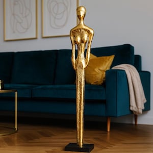 Könnte beinhalten: Eine goldfarbene, abstrakte Skulptur einer menschlichen Figur steht auf einem schwarzen Sockel. Die Skulptur befindet sich in einem Wohnzimmer mit einem tealfarbenen Sofa, goldenen Akzenten und abstrakter Kunst an der Wand. Der Raum hat eine moderne, elegante Ästhetik.