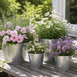 Op de afbeelding: Vier gegalvaniseerde metalen plantenbakken gevuld met kleurrijke bloemen staan op een houten tafel. De bloemen zijn onder meer roze petunia's, witte madeliefjes en paarse bloemen. De plantenbakken variëren in grootte en zijn buiten geplaatst.