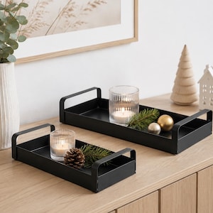 May include: Two rectangular black metal trays with integrated handles on the short ends. The trays feature a smooth matte finish and raised edges. One tray holds a ribbed glass candle holder with a lit candle, a pinecone, and greenery.