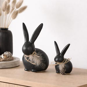 Può includere: Due statuette di coniglio in ceramica nera con lunghe orecchie e dettagli in piuma. I conigli sono decorati con spago e piume. Le statuette sono su una superficie di legno, con un vaso di fiori secchi e una candela sullo sfondo.