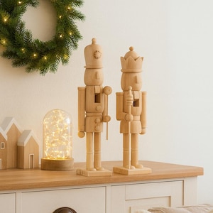 Könnte beinhalten: Zwei Holz-Nussknacker, einer mit Krone und einer mit Hut, stehen auf einer Holzoberfläche. Ein grüner Kranz mit Lichtern hängt an der Wand. Dekorative Holzhäuser und eine Glaskuppel mit Lichtern befinden sich ebenfalls auf der Oberfläche.