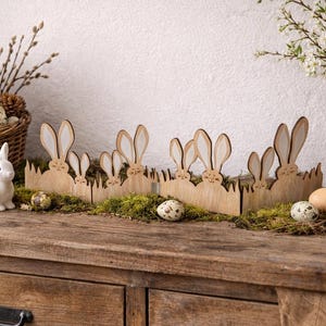 Può includere: Decorazioni a tema pasquale con un coniglietto in ceramica bianca, una recinzione in legno a forma di coniglio e un cestino di vimini con uova e fiori. Un mobile in legno rustico fa da base all'esposizione, creando una scena primaverile.