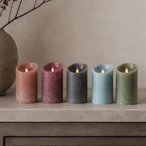 Peut inclure: Cinq bougies piliers à LED, chacune avec une lumière vacillante. Les bougies sont dans des tons de rose, violet, gris, bleu et vert. Un vase avec des branches et des baies est en arrière-plan.