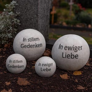 Può includere: Quattro sfere di pietra bianca di varie dimensioni, iscritte con testo nero. Due sfere recitano "In stillem Gedenken" e due "In ewiger Liebe". Le sfere sono disposte su un letto di terra marrone scuro con foglie cadute.