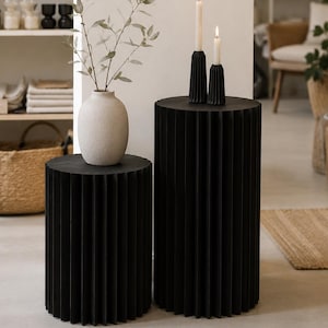 Può includere: Due tavolini cilindrici neri con una texture scanalata verticale. Un vaso beige testurizzato contenente un ramo con foglie verdi poggia sul tavolo più basso, mentre due candele bianche accese in portacandele neri si trovano su quello alto.