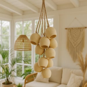 Peut inclure: Un groupe de suspensions lumineuses en forme de cloche de couleur crème, suspendues à une corde naturelle. Les lumières sont dans une pièce à la palette de couleurs neutres, avec un abat-jour tissé et un canapé en rotin.