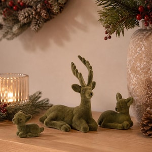 Könnte beinhalten: Ein Set aus drei olivgrünen, beflockten Hirschfiguren in ruhender Position. Der mittlere Hirsch hat ein ausgeprägtes Geweih, während die beiden kleineren Hirsche daneben liegen. Jede Figur hat eine weiche, strukturierte Oberfläche und dunkle Augen.