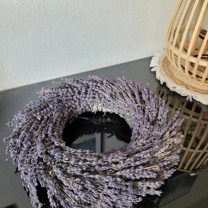 May include: A circular lavender wreath, featuring dried lavender stems arranged in a dense, symmetrical pattern. The wreath is a soft purple color and sits on a glossy black surface. A decorative wooden lamp and a framed picture are in the background.