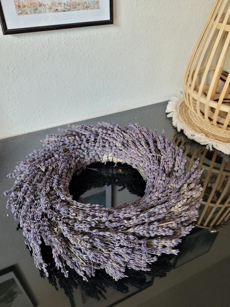May include: A circular lavender wreath, featuring dried lavender stems arranged in a dense, symmetrical pattern. The wreath is a soft purple color and sits on a glossy black surface. A decorative wooden lamp and a framed picture are in the background.