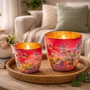 Könnte beinhalten: Zwei brennende Kerzen in dekorativen Glashaltern mit floralen Designs stehen auf einem gewebten Tablett. Die Halter haben einen roten Hintergrund mit bunten Blumen. Eine kleine Vase mit Blumen und Büchern steht ebenfalls auf dem Tisch.