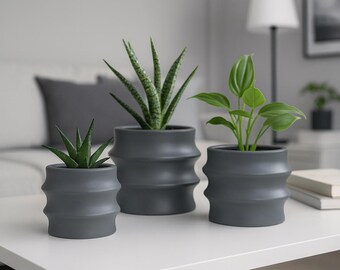 May include: Three grey ceramic plant pots with plants. The pots have a ribbed design and contain different types of greenery. The pots are arranged on a white table, with a neutral background.
