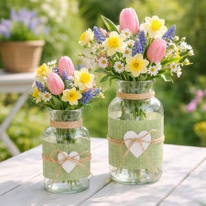 Può includere: Due vasi di vetro con fiori primaverili, tra cui tulipani rosa, narcisi gialli e giacinti blu. I vasi sono decorati con tessuto verde, spago e accenti bianchi a forma di cuore. I vasi sono su un tavolo di legno bianco.