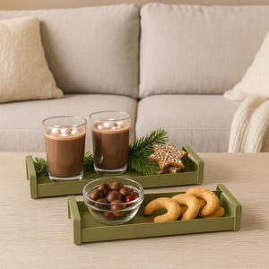 Könnte beinhalten: Zwei Gläser heiße Schokolade mit Marshmallows auf einem grünen Tablett, zusammen mit einem Lebkuchen und Tannenzweigen. Ein zweites Tablett enthält eine Schüssel mit Pralinen und Hörnchen. Die Szene spielt sich auf einem Holztisch ab.