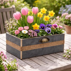 Könnte beinhalten: Ein rechteckiger Pflanzkasten aus grauem Holz mit Seildetails und einem Messinggriff, gefüllt mit rosa Tulpen, gelben Narzissen und violetten Stiefmütterchen. Der Kasten steht auf einem Holztisch.
