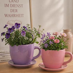 Puede incluir: Dos tazas de té de cerámica, una lavanda y otra rosa, llenas de flores coloridas. Las tazas están sobre platillos a juego. En el fondo, una impresión enmarcada con texto en alemán.