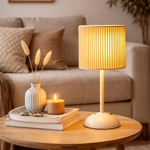 Puede incluir: Una escena interior cálida con una lámpara de mesa encendida con una base beige y una pantalla cilíndrica acanalada. Un pequeño jarrón con flores secas, una vela encendida y libros se encuentran sobre una mesa redonda de madera. Un sofá beige está en el fondo.