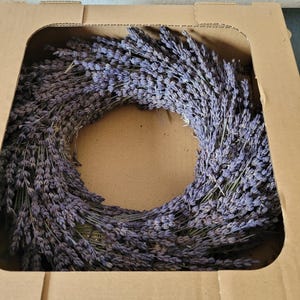 May include: A dried lavender wreath, a ring of purple flowers, sits inside a brown cardboard box. The wreath is made of many small, dried lavender stems, creating a full, circular shape. The box is open, showing the wreath.