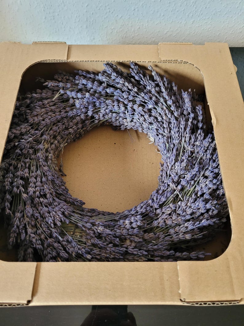 May include: A dried lavender wreath, a ring of purple flowers, sits inside a brown cardboard box. The wreath is made of many small, dried lavender stems, creating a full, circular shape. The box is open, showing the wreath.