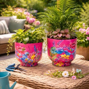 May include: Two bright pink plant pots adorned with floral patterns. Each pot contains green plants, offering a striking contrast. The pots are placed on a wicker table, with gardening tools visible, suggesting a garden setting.