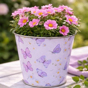 Könnte beinhalten: Ein Blumentopf aus Metall, gefüllt mit rosa Gänseblümchen mit gelben Zentren. Der Topf ist mit lila Schmetterlingen und weißen Gänseblümchen auf helllila Hintergrund verziert. Der Topf steht auf einer weißen Holzoberfläche.
