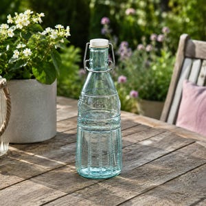 Op de afbeelding: Een heldere, lichtblauwe glazen fles met een beugelsluiting voorzien van een witte stop en een metalen draadmechanisme. De fles heeft een getextureerd oppervlak met verticale ribbeldetails rond de basis en een taps toelopende hals.