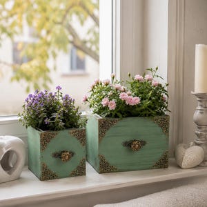 May include: Two square wooden planters in a distressed mint green finish, each featuring ornate bronze-toned metal corner brackets and a central decorative drawer pull. The smaller planter holds purple flowers, while the larger holds pink roses.