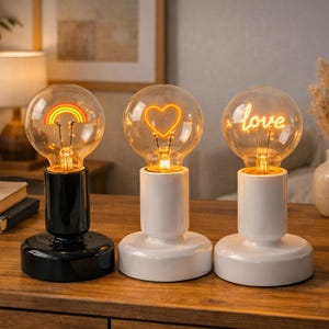 Può includere: Tre lampade decorative con lampadine e filamenti caldi e luminosi. Una lampadina presenta un arcobaleno, un'altra un cuore e la terza la parola "love". Le lampade hanno basi bianche e nere, disposte su una superficie di legno.