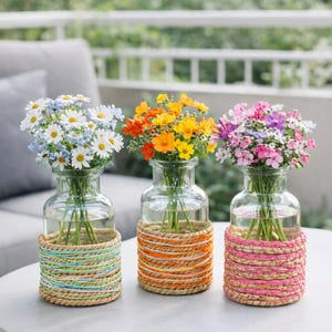 Può includere: Tre vasi di vetro con fiori colorati su un tavolo bianco. Ogni vaso ha una fascia decorativa intrecciata in diversi colori: blu e verde, arancione e rosa. I fiori includono margherite bianche, fiori arancioni e gialli e fiori rosa e viola.