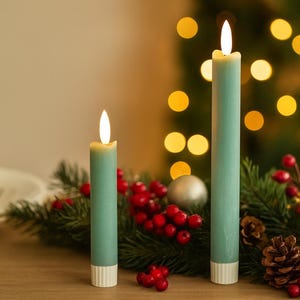 May include: Two lit teal candles with white bases are displayed on a wooden surface, surrounded by a festive arrangement of greenery, red berries, pine cones, and silver ornaments. The background features blurred golden lights.