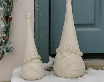 May include: Two off-white gnome statues with textured surfaces stand on a snowy step. The gnomes have long, pointed hats and detailed faces with beards. One is taller than the other. The background includes a blue door and a snowy evergreen branch.