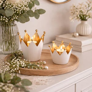 Pode incluir: Dois castiçais brancos em forma de coroa com interiores e detalhes dourados estão em uma bandeja de madeira. As velas estão acesas e a bandeja é cercada por flores e vegetação. Um toque decorativo para qualquer ambiente.