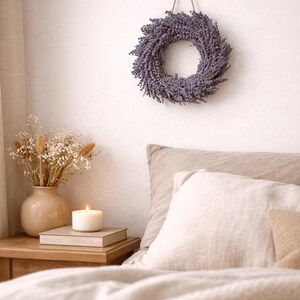 May include: A lavender wreath hangs on a white wall above a bed with beige bedding. A lit candle sits on a stack of books on a wooden nightstand next to a vase of dried flowers. The scene evokes a sense of calm and relaxation.