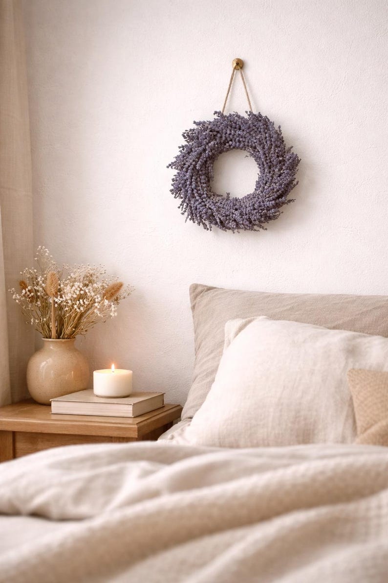 May include: A lavender wreath hangs on a white wall above a bed with beige bedding. A lit candle sits on a stack of books on a wooden nightstand next to a vase of dried flowers. The scene evokes a sense of calm and relaxation.