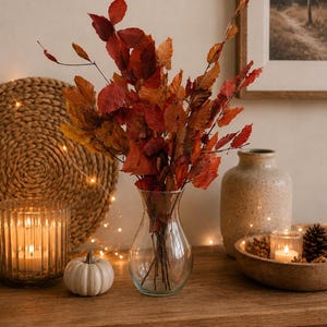 May include: A clear glass vase filled with dried autumn branches featuring vibrant red, orange, and brown leaves. The vase sits on a wooden surface alongside a small white pumpkin, a ribbed glass candle holder, and a wooden bowl with pinecones.